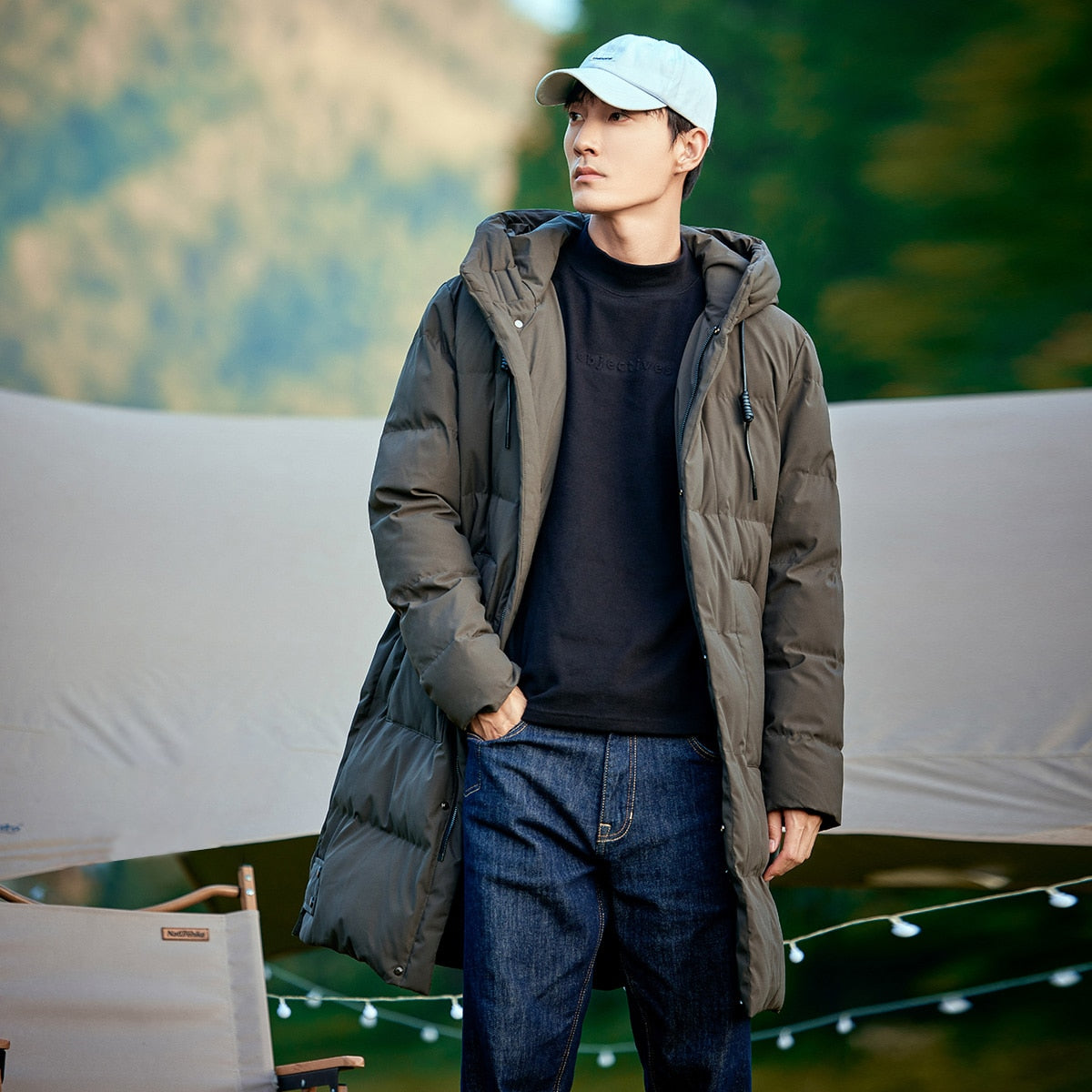 Un homme pose dans la nature. Il porte une casquette, un pull noir, un jean et un veste imperméable doudoune longue parka taupe.